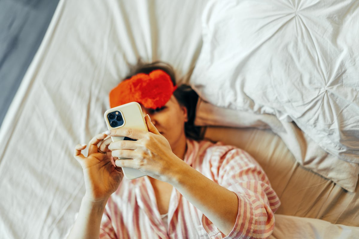 Woman in pink striped pajamas lies in bed wearing a red sleep mask while looking at her smartphone under white pillows and bedding.