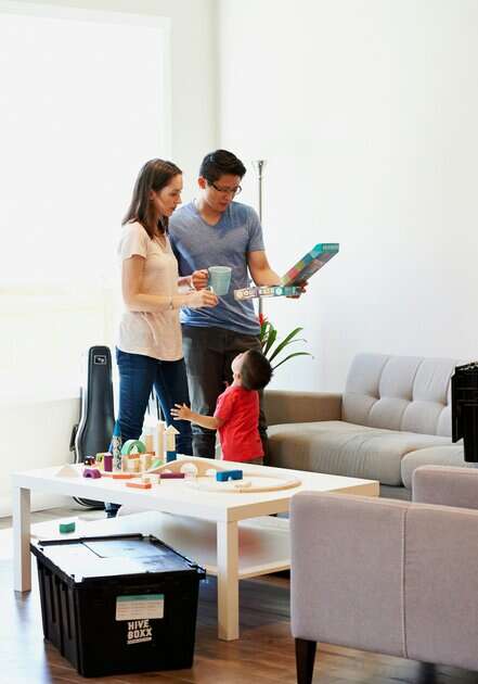 Two adults and a child stand among boxes and toys in a bright living room.