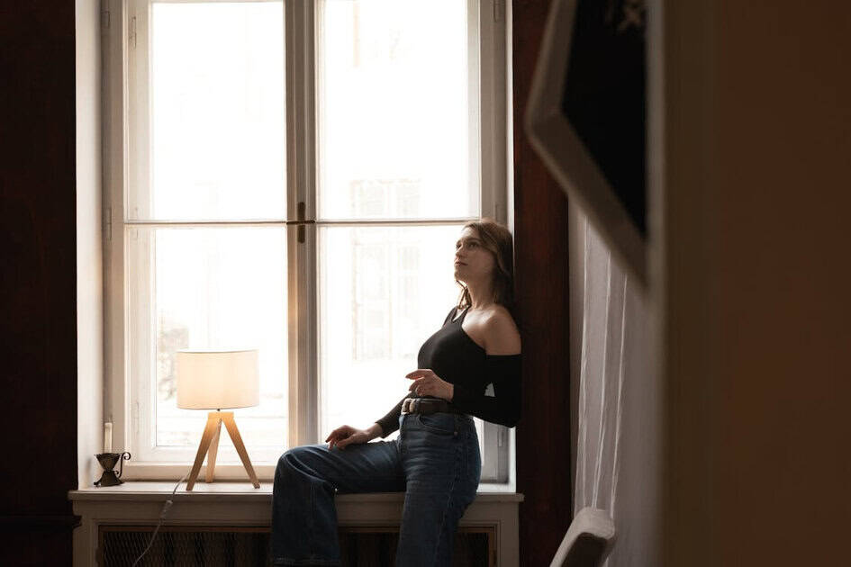 Woman sitting by a tall window in a dim room with a lit table lamp nearby