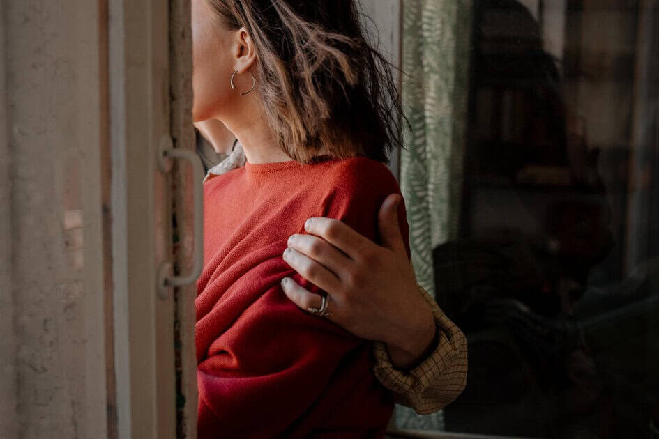 Person in a red sweater looking out a window while a hand rests on their shoulder.
