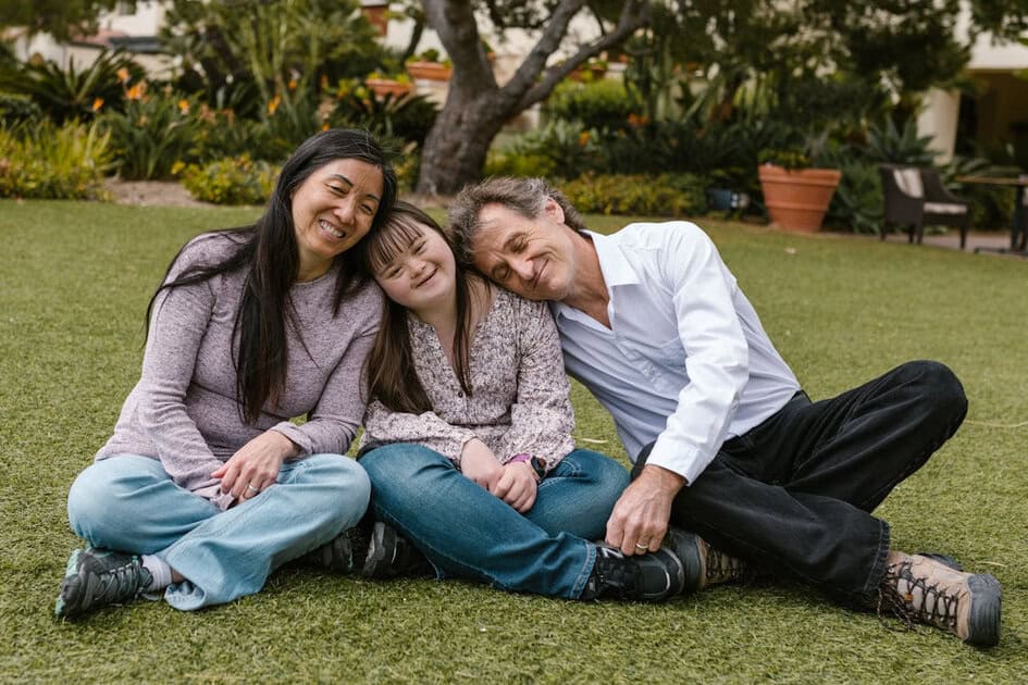 Three people sitting together on grass, smiling with their heads close in a garden setting.