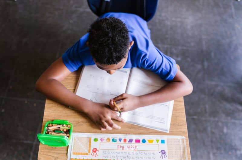 Child in a blue shirt writing in an open workbook at a desk with crayons nearby.