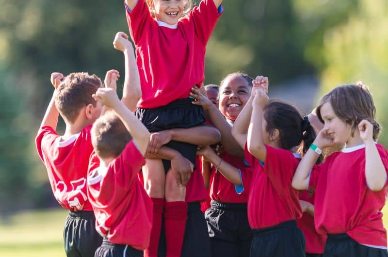 Children in red sports uniforms cheering outdoors as one child is lifted above the group.