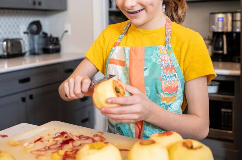 Child in a yellow shirt and patterned apron peeling an apple at a kitchen counter.