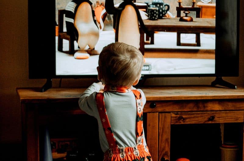 Young child in pajamas standing close to a TV showing animated penguin-like characters.