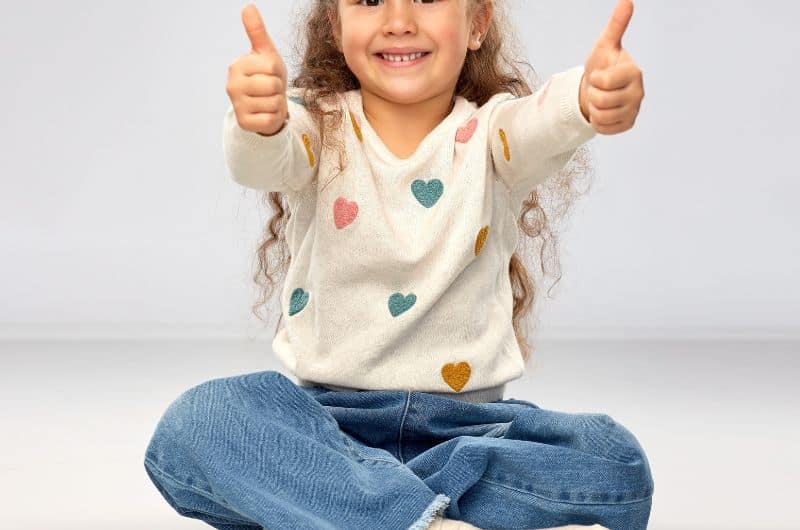 Smiling child seated cross-legged with both thumbs up, wearing a heart-patterned sweater and jeans.
