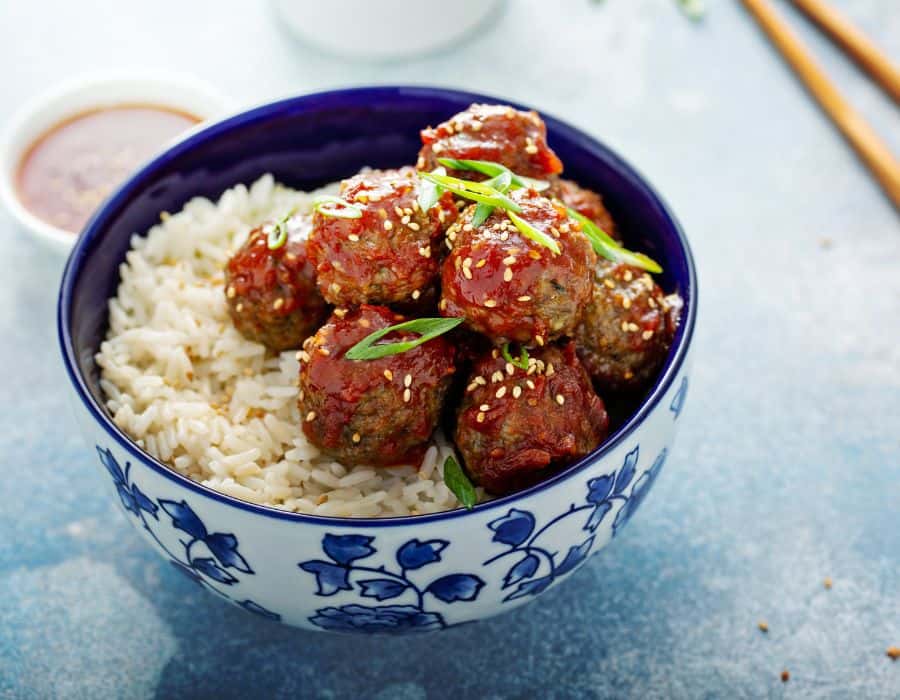 Glazed meatballs with sesame seeds and green onion served over white rice in a blue patterned bowl.