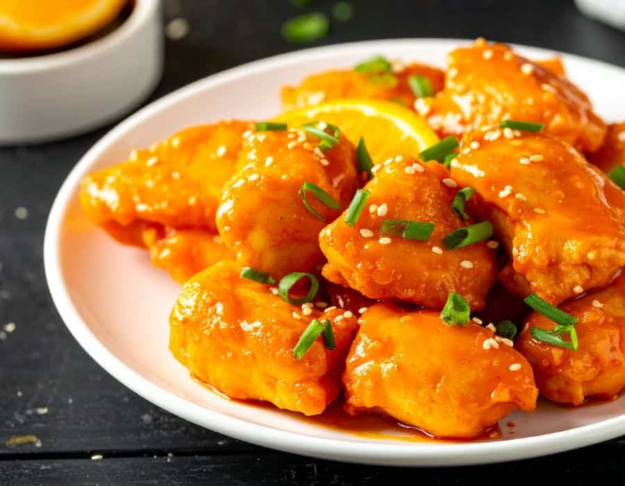 Orange-glazed chicken pieces on a white plate with sesame seeds, green onions, and an orange slice.
