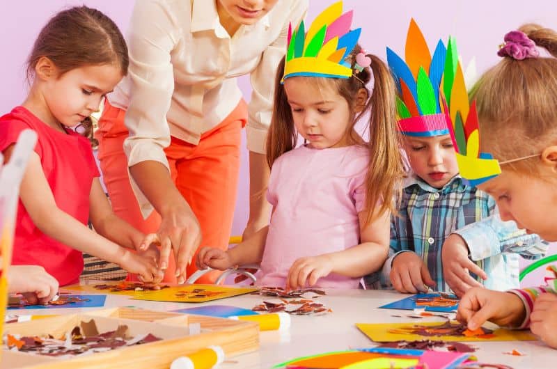 Children wearing colorful paper crowns work on crafts at a table while an adult helps.