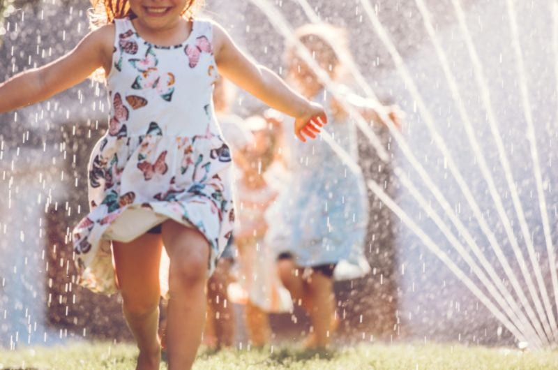Child in a butterfly-print dress running through sprinklers on grass