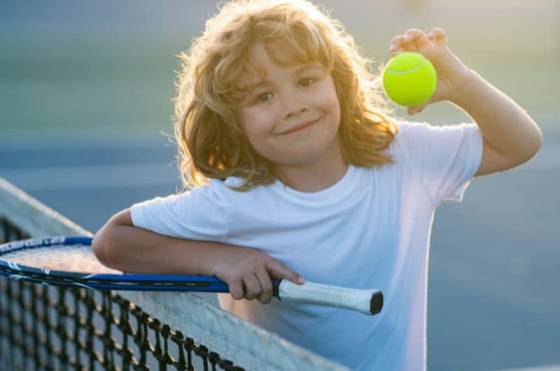Smiling child holding a tennis ball while leaning on a racket beside a court net