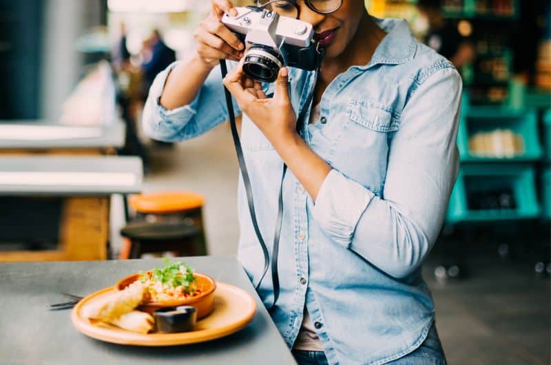 Person in a denim shirt photographing a plate of food with a camera at a table.