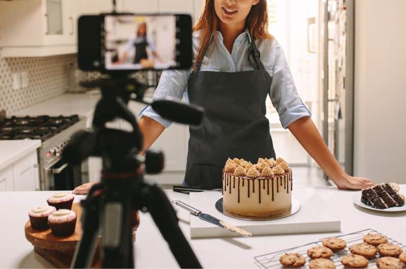 Woman in an apron recording a dessert display with a camera in a kitchen
