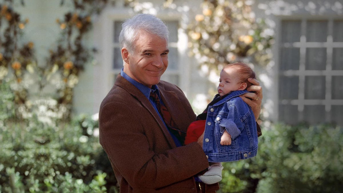 Smiling gray-haired man in a brown jacket holding a baby in a denim jacket outdoors