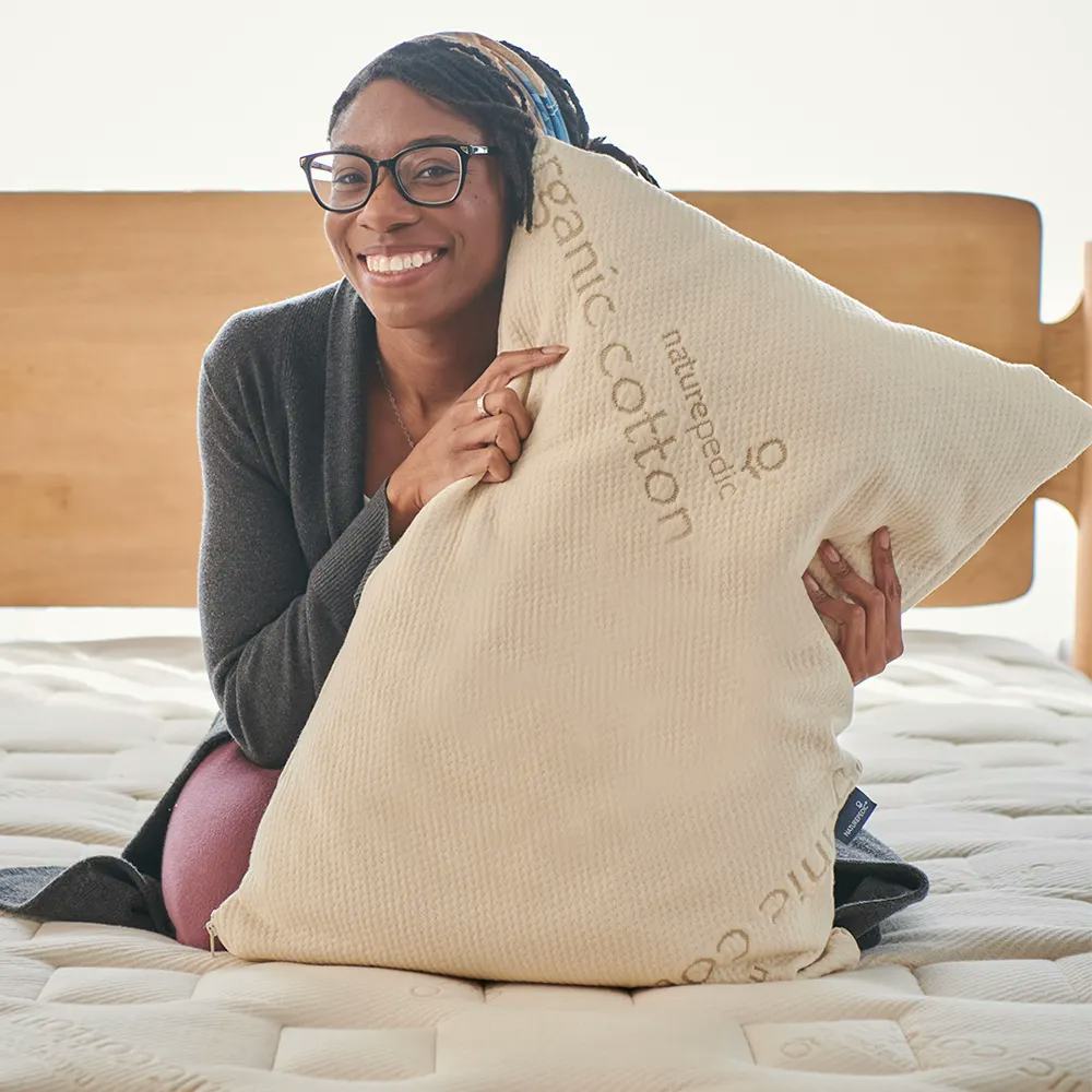 Smiling woman with glasses sits on a bed holding a large cream pillow printed with organic cotton text.