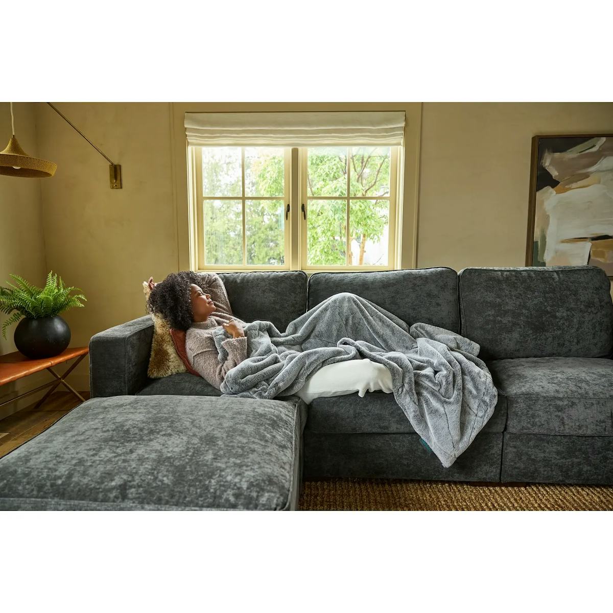 Woman resting under a gray blanket on a dark sectional sofa in a sunlit living room.