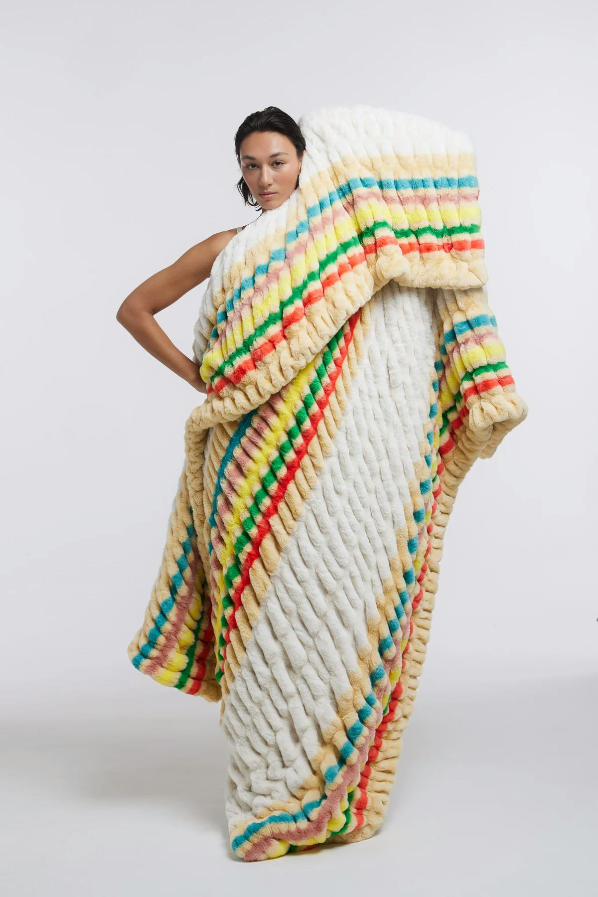 Woman wrapped in an oversized cream plush blanket with bright multicolor stripes on a plain studio backdrop.