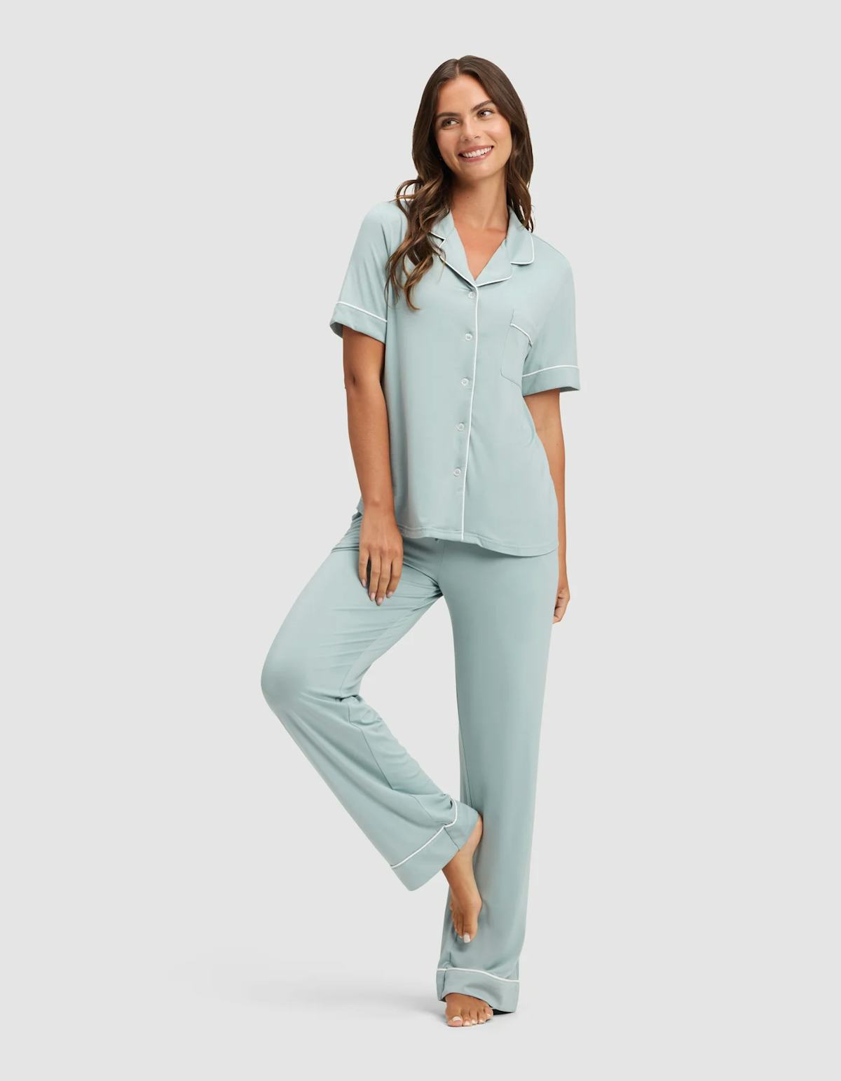Smiling woman poses barefoot in pale blue short-sleeve pajamas on a plain gray background.