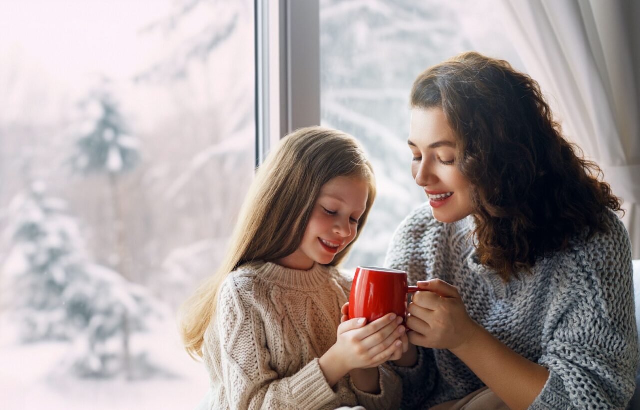 Woman and young girl in sweaters smiling together by a window while holding a red mug.