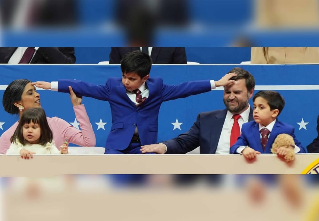 Boy in a blue suit stretches his arms between seated adults and children in a blue-railed venue.