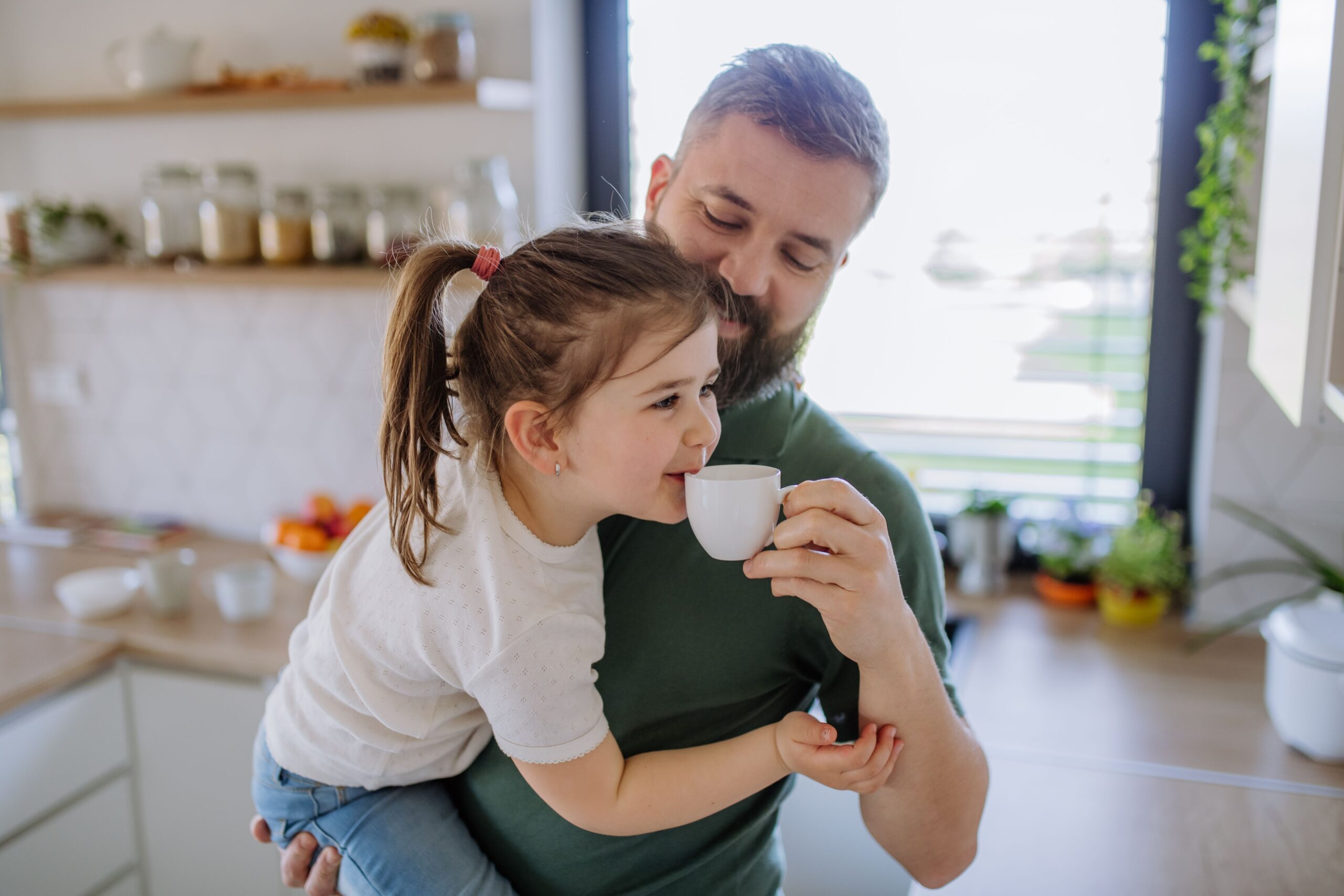 Adult man holding a young child as the child sips from a small white cup in a bright kitchen.