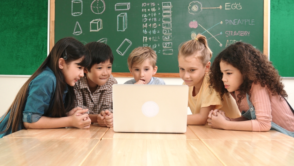 Five children gather around a laptop in front of a chalkboard with drawings and fruit labels.