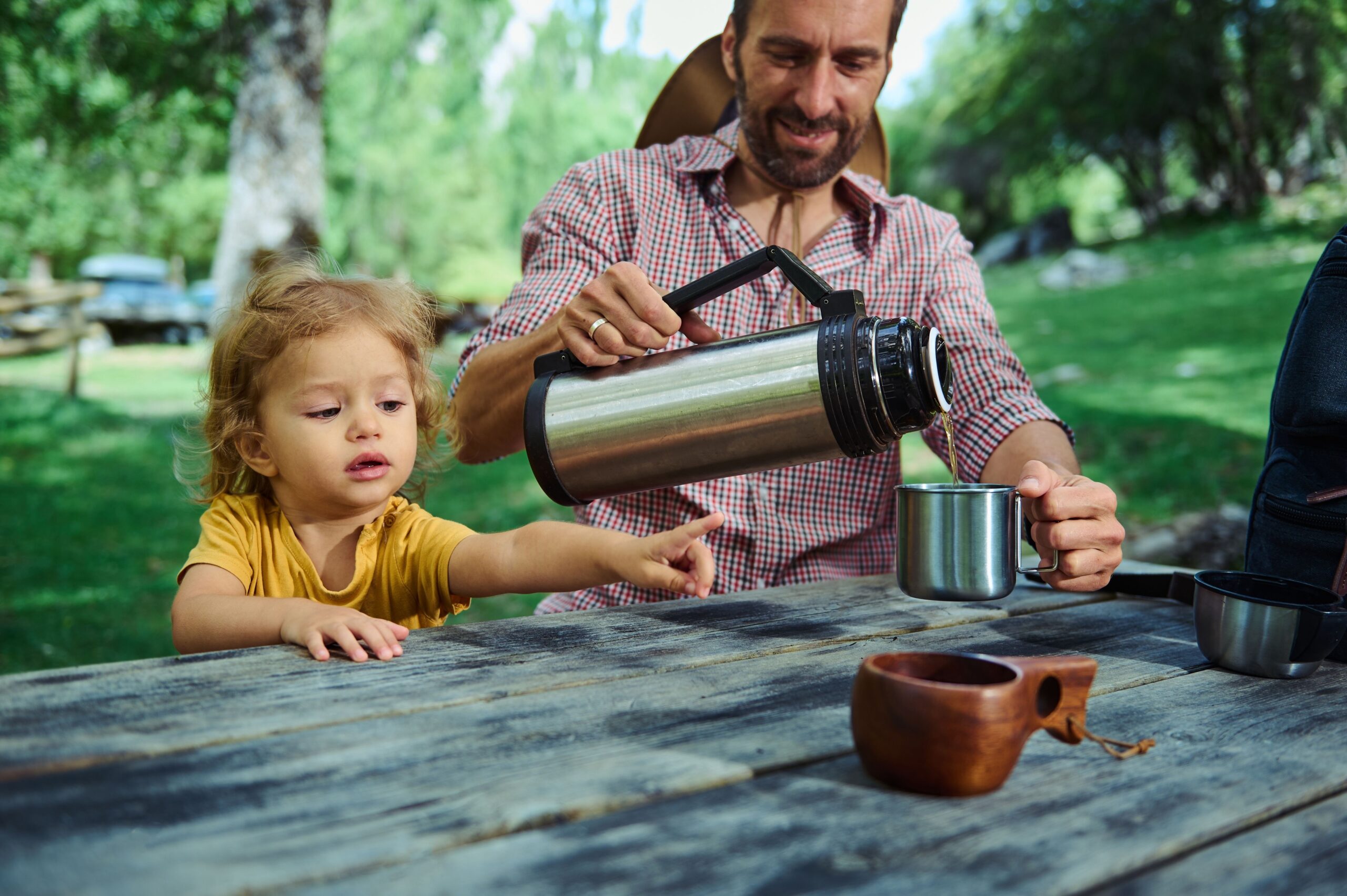 Adult pouring a drink into a metal cup while a young child reaches across a picnic table