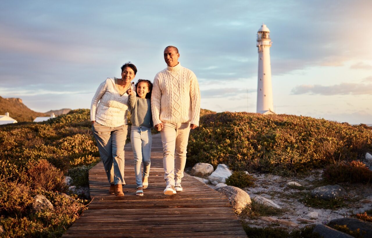 Two adults and a child walk on a wooden path near a lighthouse at sunset.