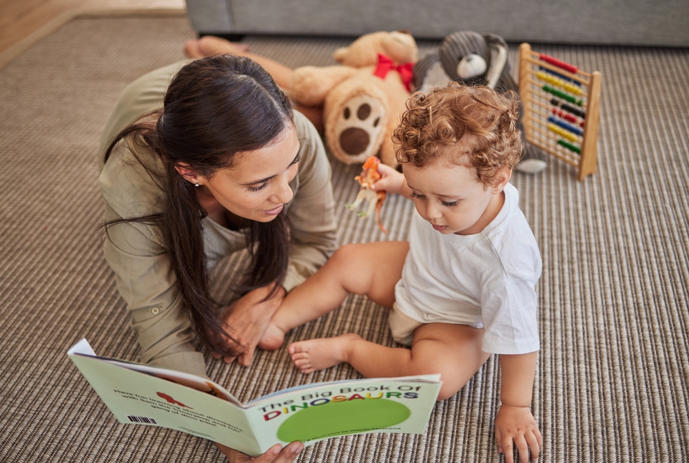 Adult woman reads a dinosaur book with a young child seated on a carpet with toys nearby.