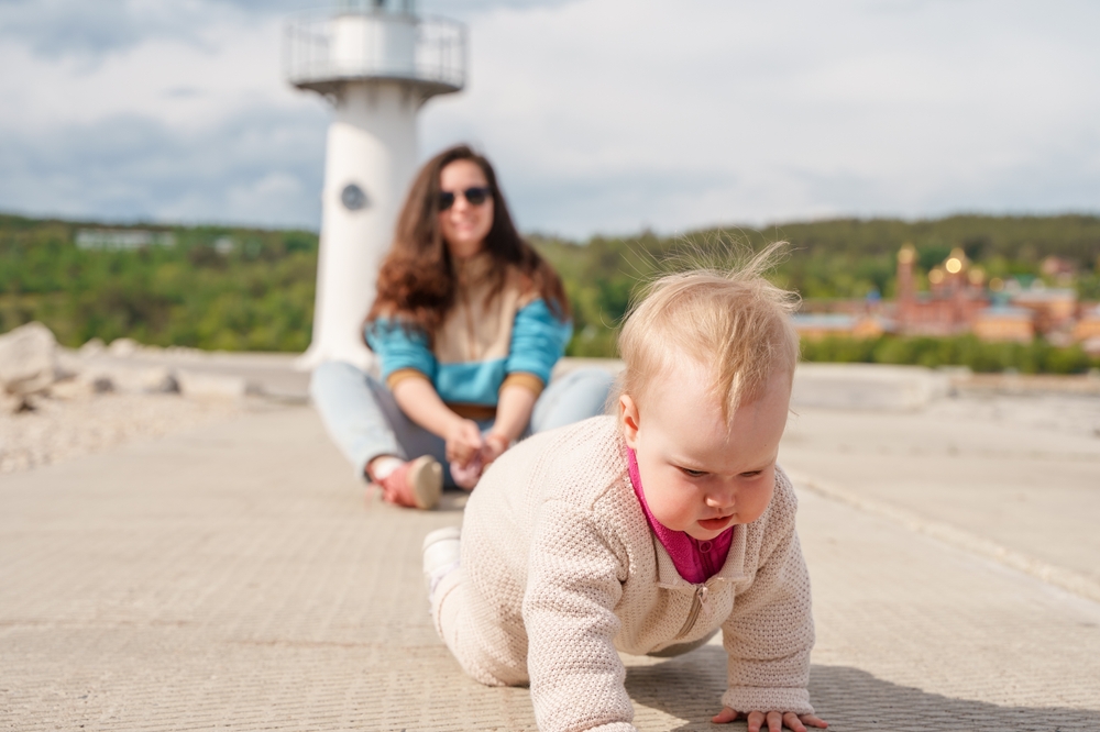 Baby crawling on a paved surface with a seated adult and white lighthouse-like tower blurred behind.