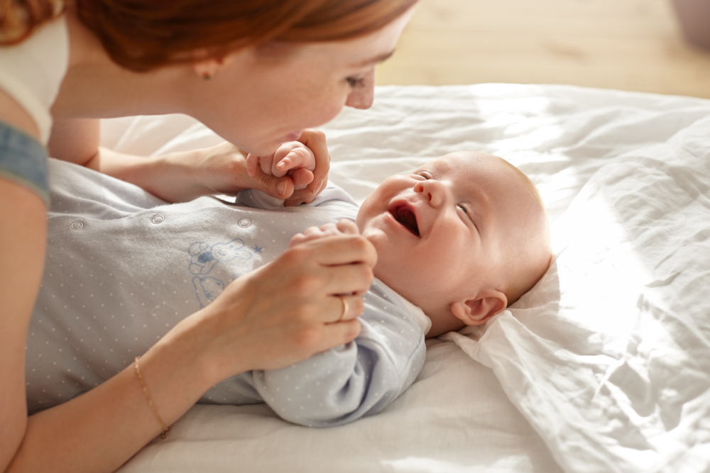 Adult woman smiling at a baby lying on a white bed and holding the baby's hands.