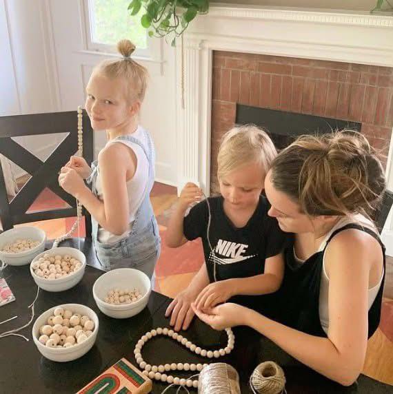Adult and two children stringing wooden beads at a table with bowls of beads.
