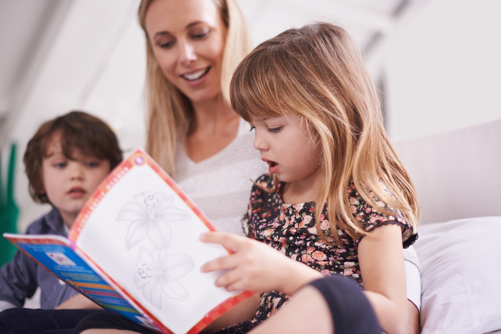 Young girl holds an open book while a woman and another child sit nearby indoors.
