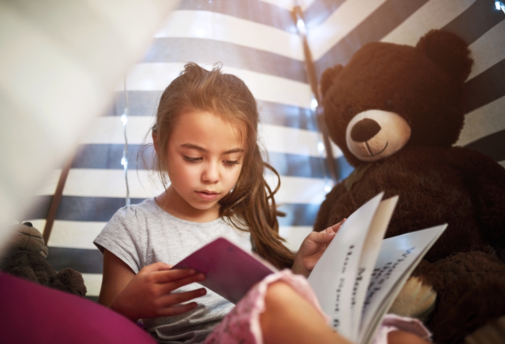 Young girl reading a book beside a large teddy bear under striped fabric and soft lights