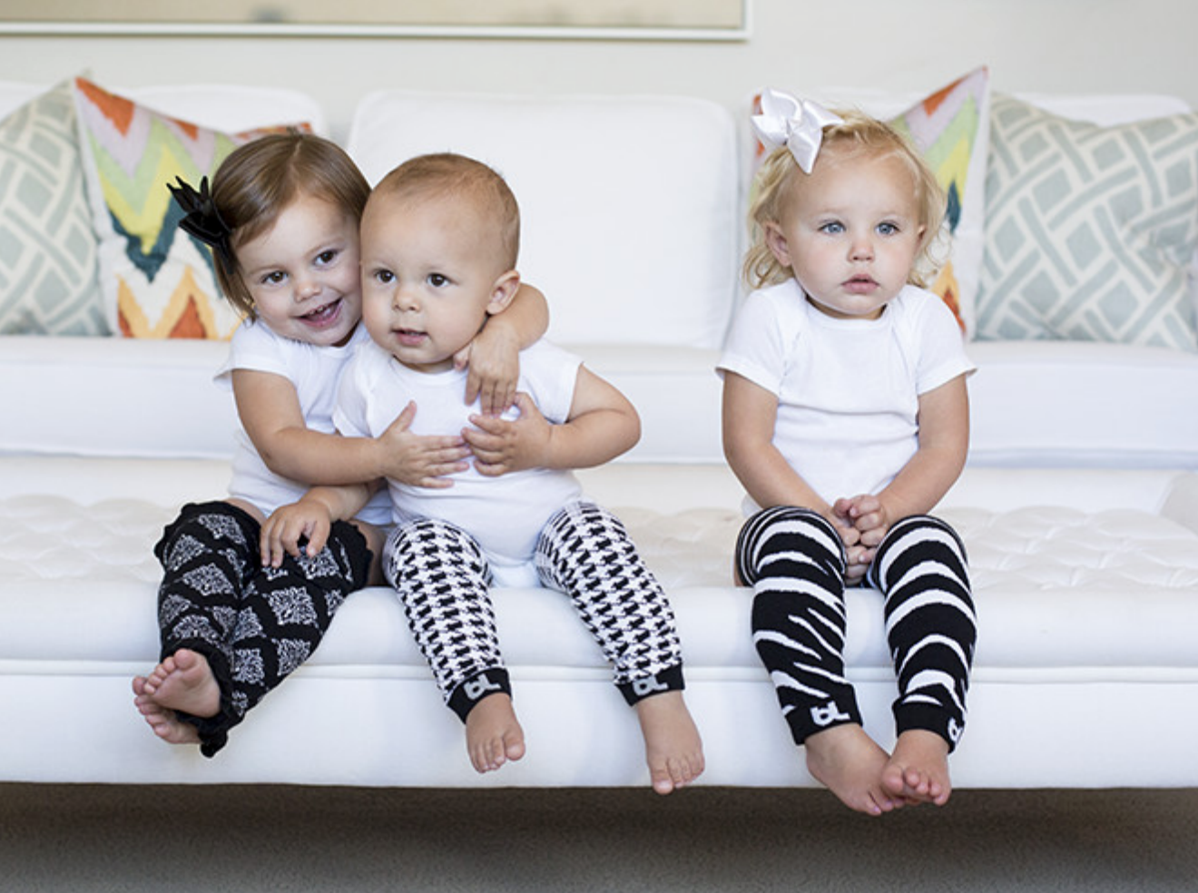 Three toddlers in white shirts and patterned leggings sitting on a white sofa.