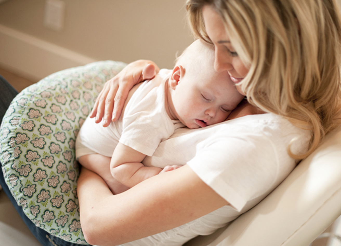 Light-haired woman cradling a sleeping baby against her chest on a patterned cushion.