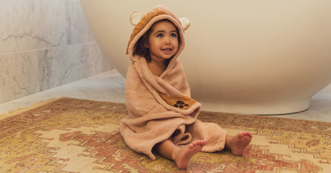 Young child wrapped in a hooded towel sitting on a patterned rug beside a bathtub.