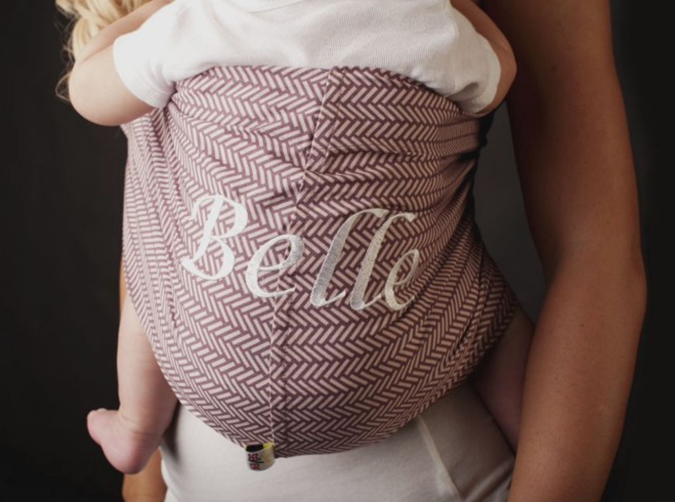 Baby carried in a patterned fabric wrap with the word Belle embroidered across the front