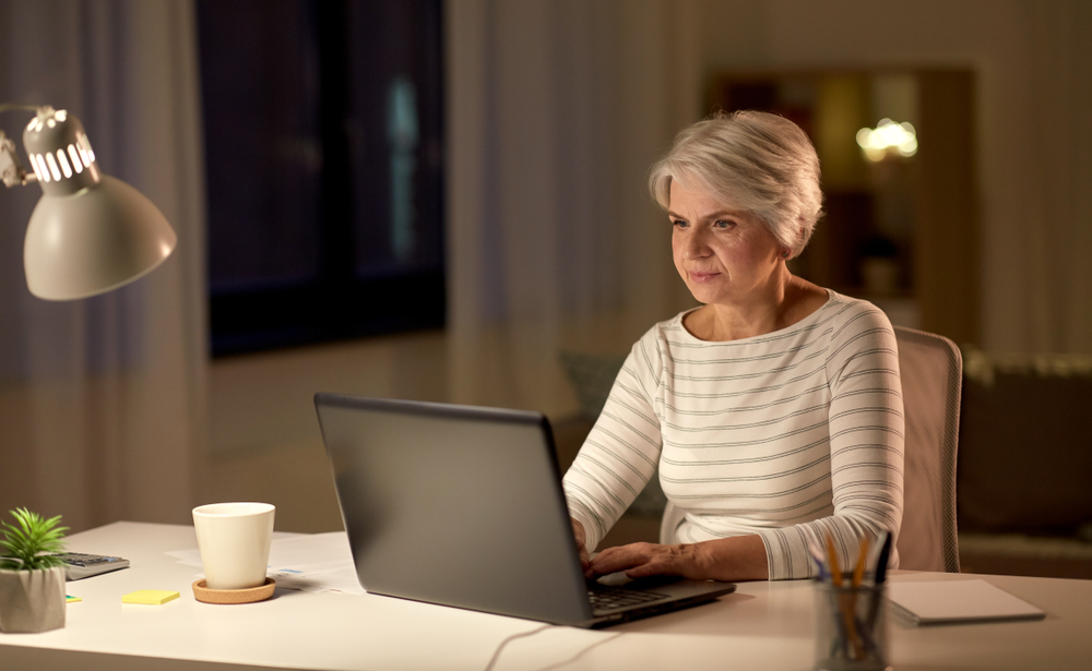 Gray-haired woman using a laptop at a desk under a lamp in a dim room