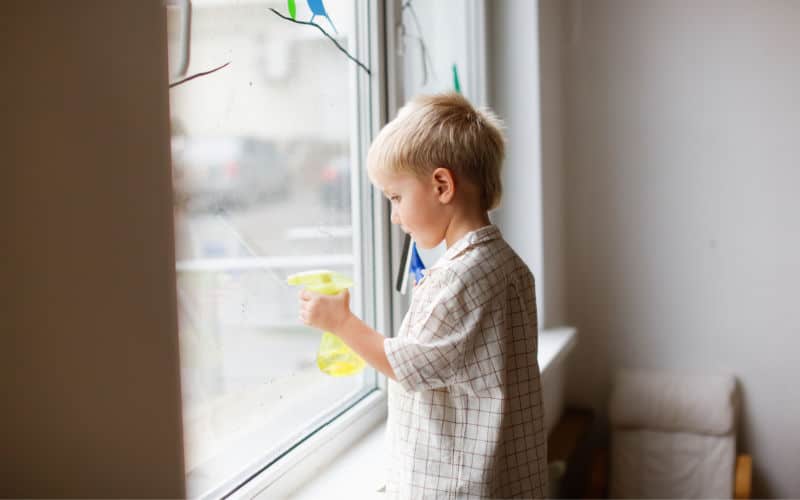 Young blond child holding a yellow cloth while standing beside a large window indoors.