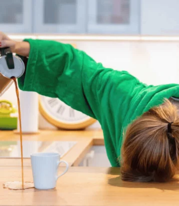 Person in a green top slumped on a kitchen counter beside a white mug and spilled coffee.