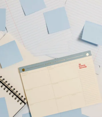 Weekly planner on a desk with blue sticky notes, lined paper, and a spiral notebook nearby