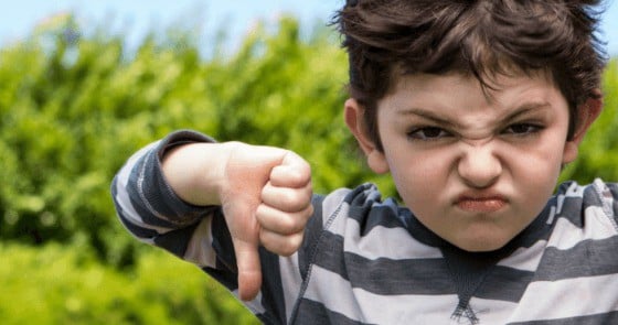 Young boy in a striped shirt frowns and gives a thumbs-down outdoors.