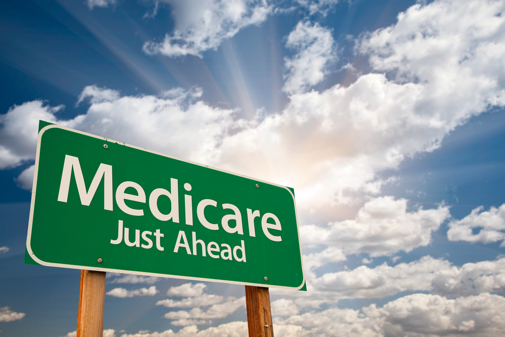 Green roadside sign reading "Medicare Just Ahead" against a bright cloudy sky.