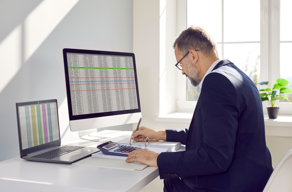 Man at a desk using a calculator beside laptop and desktop screens showing spreadsheets