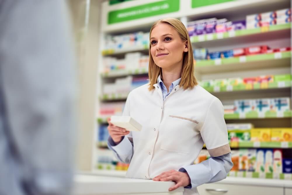 Woman in a white coat holding a box at a pharmacy counter with shelves behind her.