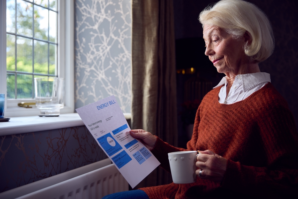 Older woman holding an energy bill and a mug while sitting beside a window and radiator.