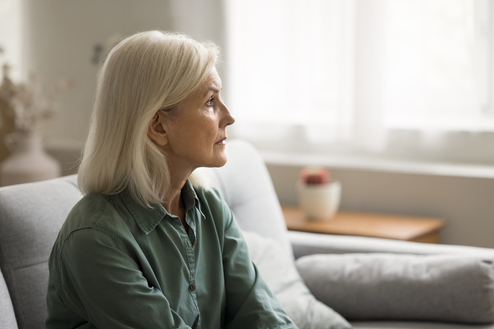 Older woman with gray hair sitting on a sofa and looking toward a bright window
