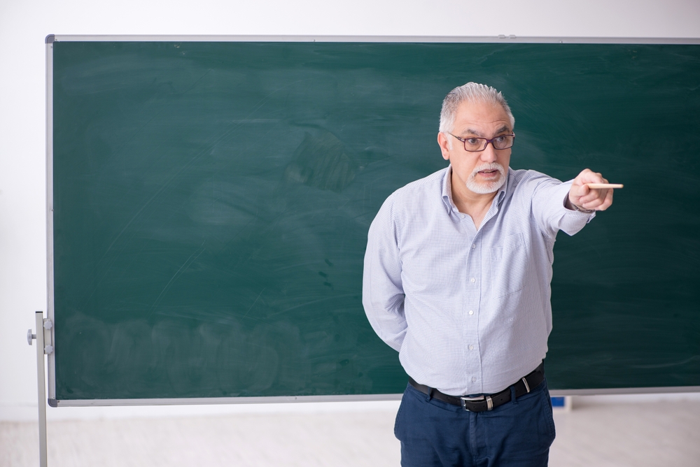 Older man with glasses points with chalk while standing in front of a large green chalkboard.