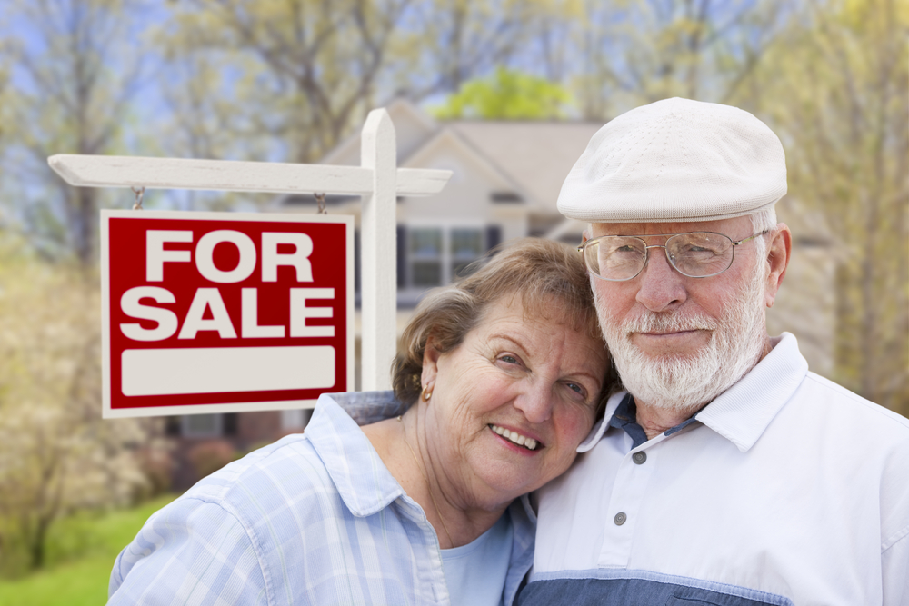 Older couple standing beside a red and white "FOR SALE" sign in front of a house
